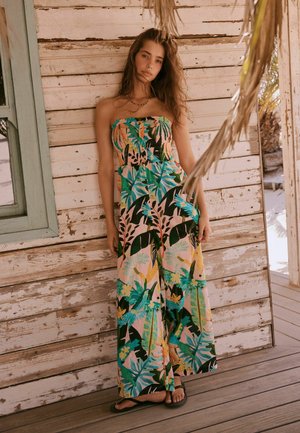 Junge Frau in einem trägerlosen, weit geschnittenen Overall mit buntem tropischem Blatt- und Vogelmuster, stehend vor einer verwitterten Holzwand.