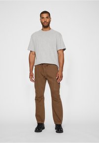 Lichtgrijs shirt met korte mouwen, gecombineerd met bruine cargobroek met zijzakken, gedragen met zwarte schoenen. Eenvoudig ontwerp met losse pasvorm.