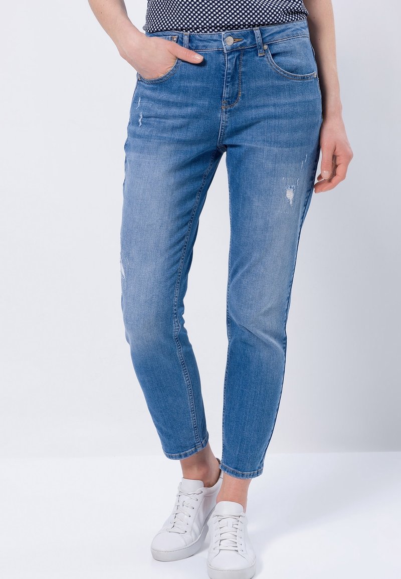 Personne portant un jean slim bleu légèrement usé avec un ourlet roulé et des baskets blanches, debout, une main dans la poche.