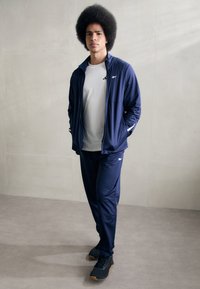 Chándal atlético en color navy, elaborado con una tela suave, que incluye una chaqueta con cremallera y detalles en blanco, combinada con pantalones ajustados a juego y zapatos negros.