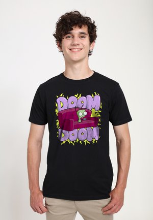 Jeune homme souriant portant un t-shirt noir avec un extraterrestre de dessin animé sur un canapé violet avec le texte "DOOM DOOM" entouré de formes d'éclairs jaunes.