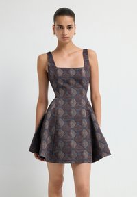 Femme portant une robe mini sans manches, à motifs, évasée avec un décolleté carré et des boucles d'oreilles en or, se tenant devant un fond blanc.