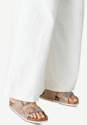 Füße in metallisch roségoldfarbenen Sandalen mit Schnallen, kombiniert mit weißen Hosen mit weitem Bein auf hellem Hintergrund.
