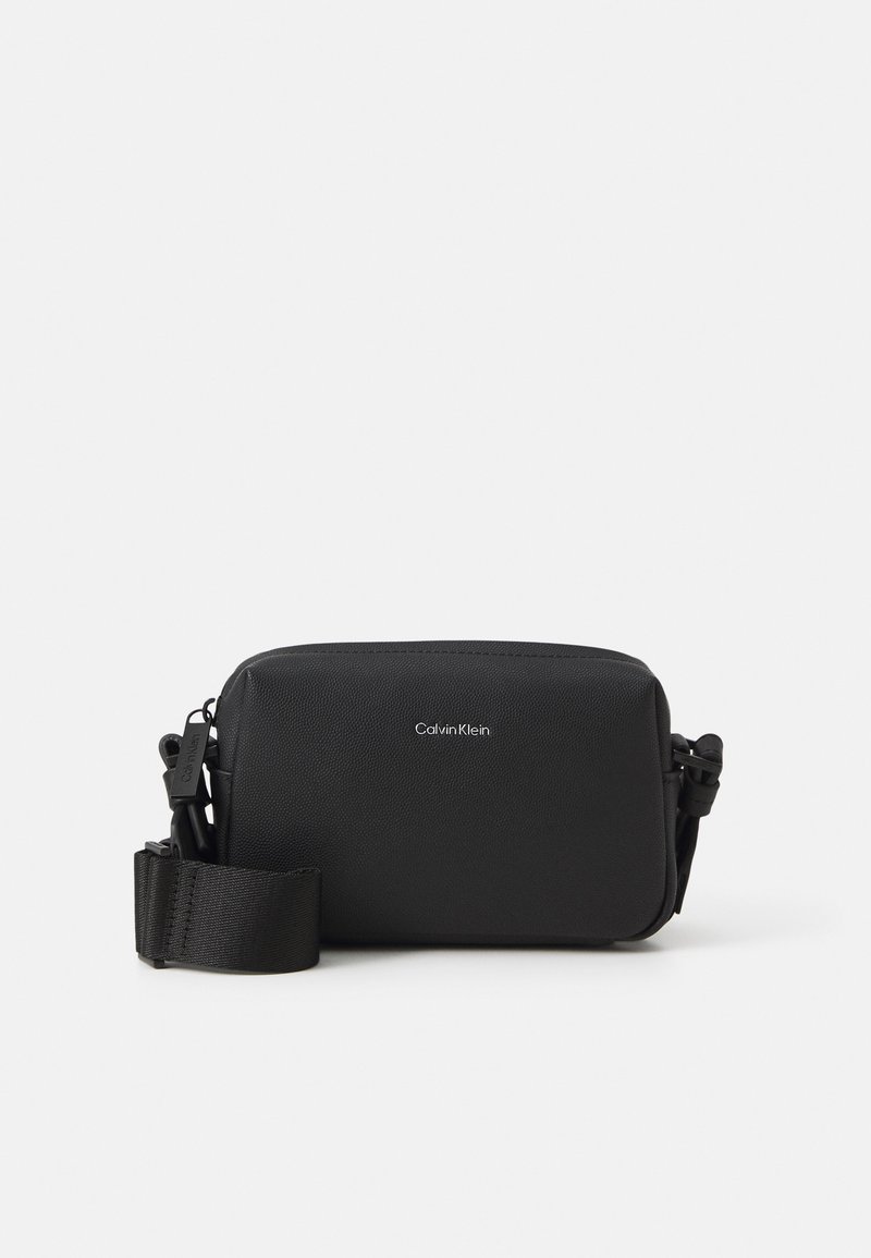 Sac en bandoulière en cuir noir avec une finition texturée, fermeture zippée, sangle réglable et logo Calvin Klein à l'avant.