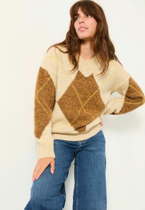 V-Ausschnitt-Pullover mit beigem Grund und braunen sowie gelben Rautenmuster. Weicher Stoff, lockere Passform und gerippte Bündchen. Kombiniert mit blauen Jeans.