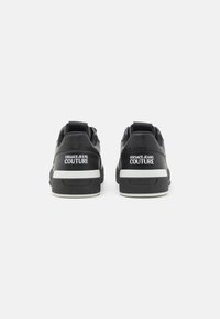 Sorte lærersneakers med hvite såler, med "VERSACE JEANS COUTURE" merkevare på hælen og teksturerte detaljer. Enkel og moderne design.