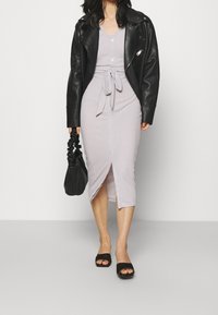 Hellviolettes geripptes Midikleid mit einem Schlitz vorne, Taillendetail zum Binden und weißen Knöpfen, kombiniert mit einer schwarzen Lederjacke und einer schwarzen Handtasche.