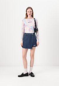 Lagani, dugih rukava majica s pastelno tie-dye uzorkom, uparena s plavom traper suknjom na preklop koja sadrži logo i detalj s bočnim dugmetom.
