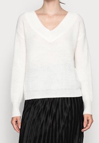 Maglione bianco lavorato a maglia con profondo scollo a V, maniche lunghe e orlo a coste. La texture è leggera e ariosa, complementando la forma dell'indumento.