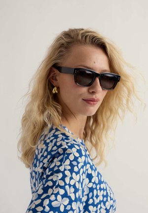 Blonde vrouw met zwarte rechthoekige zonnebril, gouden oorringen en een blauwe top met een wit bloemenpatroon, kijkt lichtjes opzij.