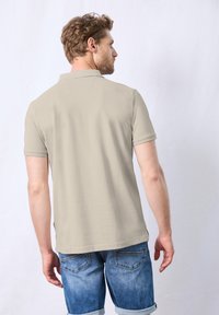 Béžové polo tričko s krátkým rukávem, s klasickým límečkem, v kombinaci s vybledlými modrými džínovými šortkami. Jednoduchý design, hladká textura.