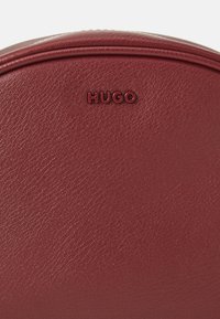 Rød teksturert læroverflate med preget "HUGO"-logo i matchende farge nær øverste midtpunkt, buet kantdetalj over.