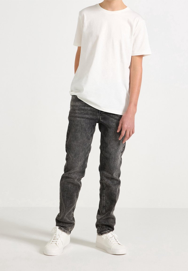 Personne portant un t-shirt blanc uni à manches courtes, un jean noir délavé et des baskets blanches, debout devant un fond blanc uni.