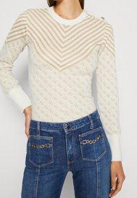 Heller Creme-Pullover mit goldfarbenem Chevron-Muster und strukturierter Strickoptik, kombiniert mit hochtaillierten blauen Jeans, die dekorative goldene Kettenakzente an den Taschen aufweisen.