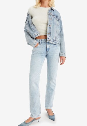 Veste en denim bleu clair et jeans taille haute, associés à un crop top crème. Mules bleues à talons fins. Texture lisse, coupe décontractée.