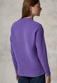 Pull à côtes violet avec des manches longues, col rond et fentes sur les côtés ; présenté sur une silhouette contre un fond texturé clair.