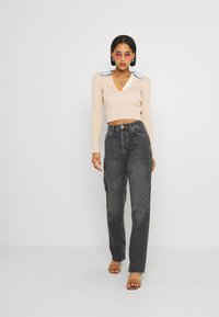 Sweater de malha em bege com colarinho azul claro, combinado com jeans escuros de cintura alta; modelo usa sandálias transparentes e óculos de sol cor-de-rosa.
