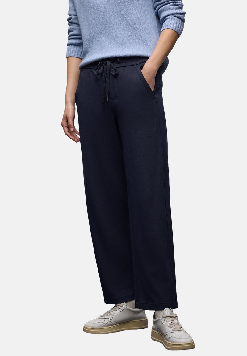 Marineblaue Sweatpants mit weitem Bein, Kordelzug in der Taille, seitlichen Taschen und einer glatten Textur, kombiniert mit hellgrauen Sneakers.