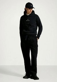 Sweatshirt preta com bolso frontal e cordões, combinada com calças cargo pretas. Usada com um boné combinando e ténis pretos.