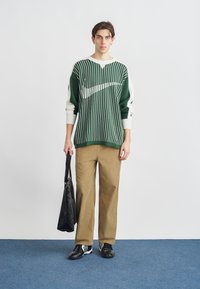 Zöld-fehér csíkos laza fazonú pulóver, khaki kordbársony nadrág, fekete táska a kezében, fekete tornacipőben. Egyszerű dizájn, hétköznapi stílus.