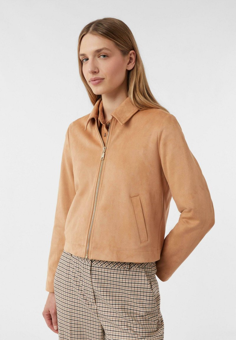 Donna che indossa una giacca in suede color beige con zip e pantaloni a quadri a vita alta, in piedi con una mano dietro la schiena.
