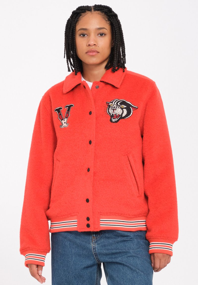 Veste en polaire rouge avec un col, boutons pression noirs, poches latérales et poignets rayés. Présente des patches brodés d'un tigre et de la lettre "V".