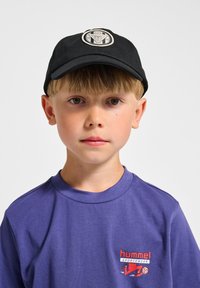 Svart keps med logotyp på framsidan, ihop med en blå skjorta med en röd grafisk design. Kort hår och neutral min.