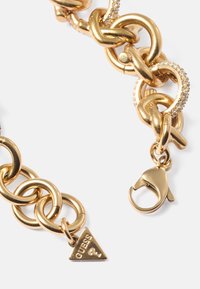Pulseira de corrente em ouro com elos entrelaçados, um design texturizado, detalhes brilhantes e um charm triangular "Guess" com fecho tipo lagosta.