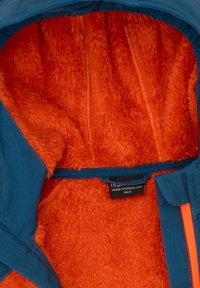 Blauwe lijnenjack met een oranje pluizige binnenkant. Kenmerken zijn onder andere een ritssluiting en een label. Zachte texturen zijn overal zichtbaar.