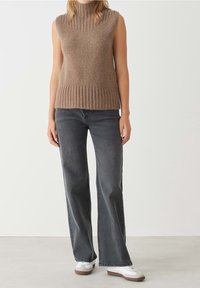 Ärmelloser brauner Strickpullover mit Rippstruktur, kombiniert mit dunkelgrauen Jeans mit weitem Bein und weißen Sneakers. Weicher Stoff mit figurbetontem Schnitt.