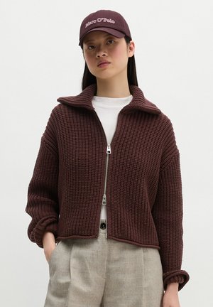 Femme portant un pull zippé en maille bordeaux, une chemise blanche, un pantalon beige et une casquette de baseball bordeaux assortie sur un fond uni.
