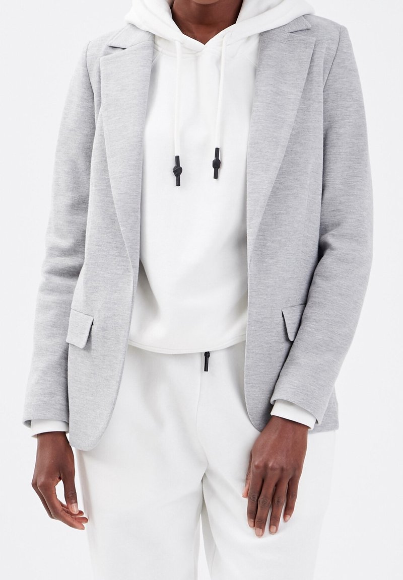 Personne portant un blazer gris clair par-dessus un sweat à capuche blanc et un pantalon blanc sur un fond blanc uni.