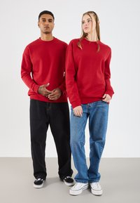 Twee personen dragen bijpassende rode crewneck sweatshirts. De een combineert het met zwarte losse broek en sneakers; de ander met blauwe jeans en witte schoenen.