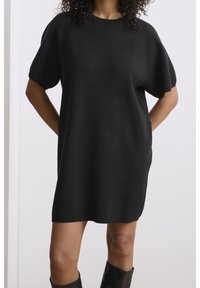 Femme portant une robe-pull noire ample à manches courtes, debout avec les mains derrière le dos sur un fond uni.