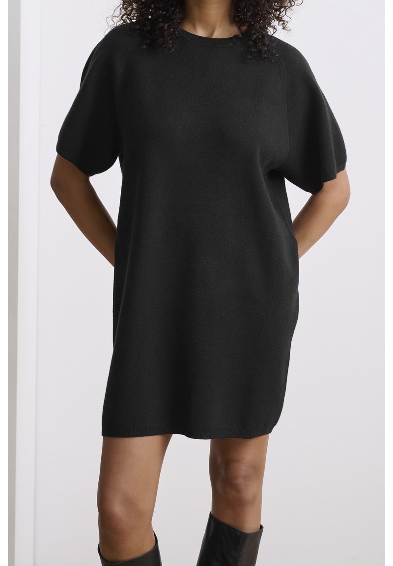 Femme portant une robe-pull noire ample à manches courtes, debout avec les mains derrière le dos sur un fond uni.