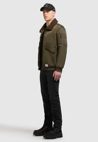 Jachetă bomber verde măsliniu cu guler din blană maro; are buzunare cu fermoar, manșete ribbate și un logo pe patch. Blugi negri și bocanci masivi negri.