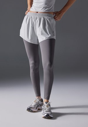 Personne portant un short de sport blanc par-dessus des leggings gris et des chaussures de course blanches, debout devant un fond gris.