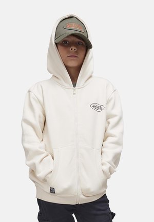 Jeune personne portant un sweat à capuche zippé crème avec le logo "Von Dutch" et une casquette verte olive avec une broderie orange "Von Dutch".