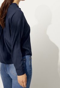 Marineblaue Bluse mit langen Ärmeln, hohem Ausschnitt und gerafftem Saum, kombiniert mit eng geschnittenen, hellblauen Jeans. Glatte Textur.