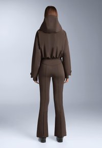 Hnedý dvojdielny outfit so priliehavou bundou s kapucňou a rozšírenými nohavicami, obidva kusy s jemnými švovými detailmi a hladkou textúrou.