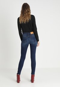 Pantalones vaqueros ajustados de cintura alta en denim azul oscuro. Con un parche de cuero marrón en la parte trasera, costuras laterales y dos bolsillos traseros.