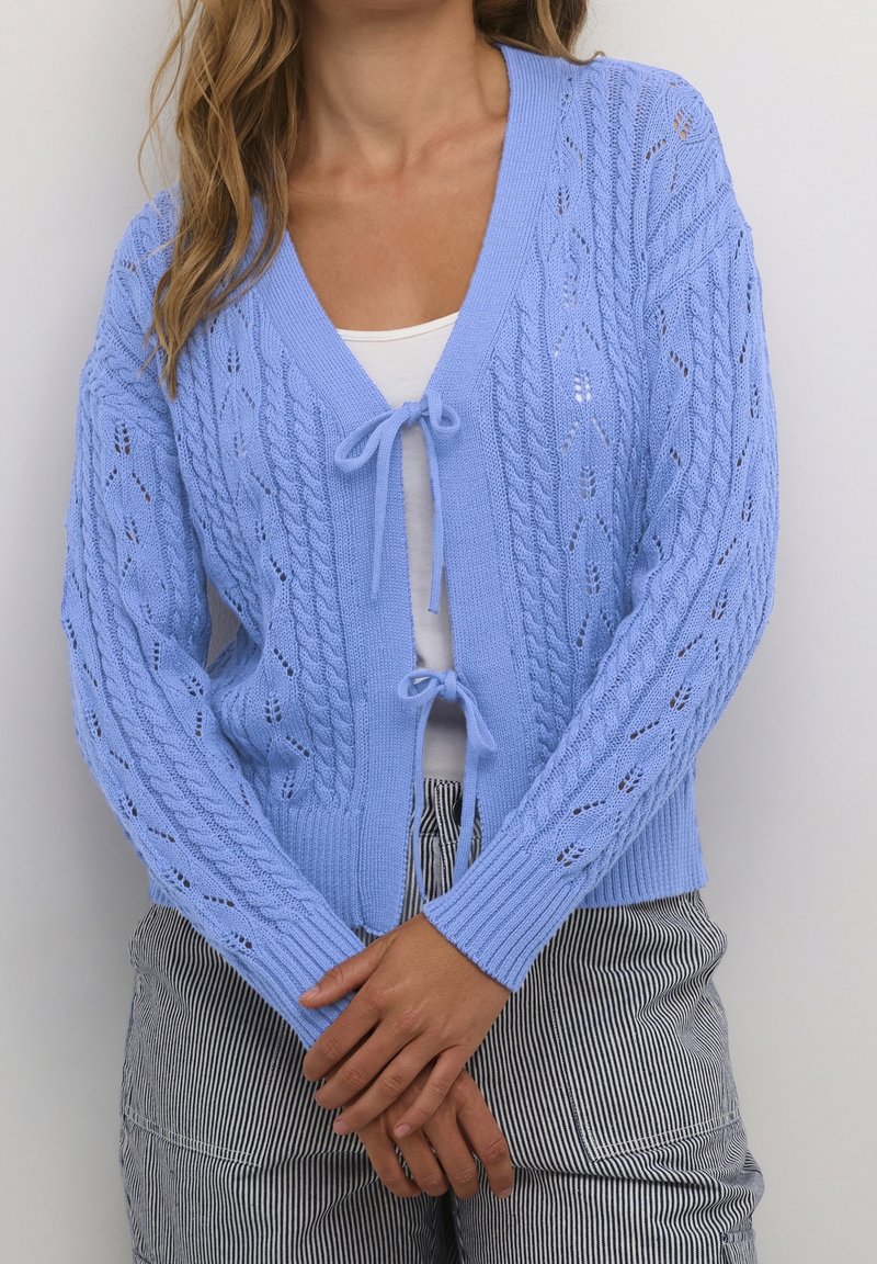 Jasnoniebieski dziany kardigan z dekoltem w serek, zapinany na wiązanie, ozdobiony wzorem w warkocze. Długie rękawy i ściągacze przy mankietach dodają tekstury.