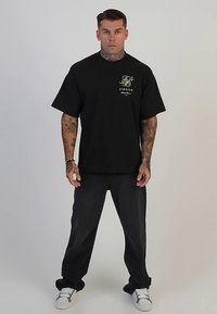 Crna majica kratkih rukava sa zlatnim logotipom na lijevoj strani prsa, uparen s širokim crnim hlačama i bijelim tenisicama. Neutralna pozadina.