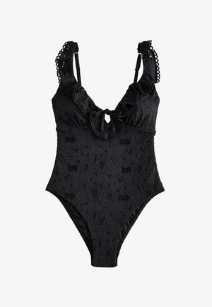 Frau mit langem dunklem Haar in einem schwarzen Spitzenbadeanzug posiert mit einem erhobenen Arm zwischen blühenden Hortensiensträuchern mit blauen und rosa Blumen.