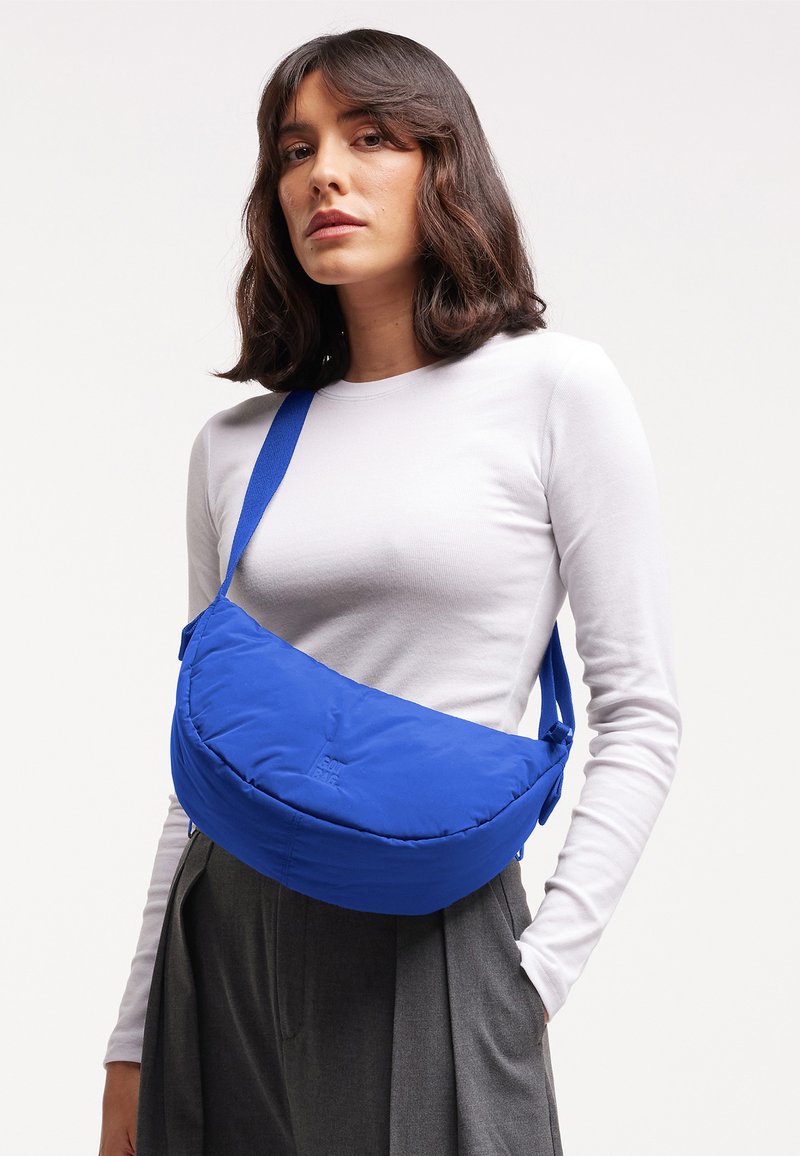 Kvinde med skulderlangt mørkt hår iført en hvid langærmet skjorte og grå bukser, der bærer en blå crossbody-taske.