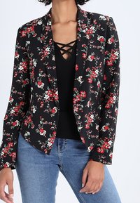 Blazer noir à motif floral avec des fleurs rouges et blanches, tissu léger, revers crantés, devant ouvert, porté sur un haut noir, associé à un jean bleu.