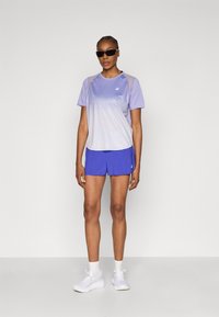 Μωβ αθλητική μπλούζα με διαβαθμισμένο σχέδιο και λογότυπο, σε συνδυασμό με μπλε σορτς. Το μοντέλο φοράει μαύρα γυαλιά ηλίου και λευκά αθλητικά παπούτσια.