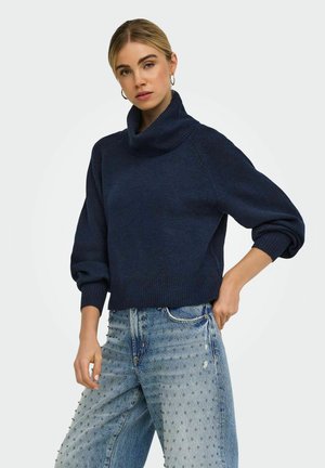 Femme aux cheveux blonds portant un pull à col roulé bleu marine et un jean bleu clair avec de petits clous, posant avec une main dans la poche.