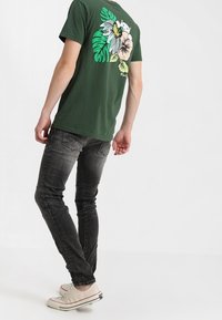 Camiseta de algodón verde con un diseño floral en la parte posterior. Combinada con jeans gris oscuro y zapatillas claras. Ajuste sencillo y casual.