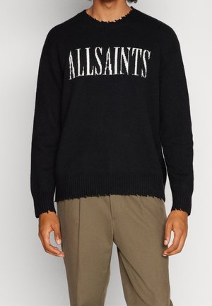 Homme portant un pull noir avec « ALLSAINTS » en lettres blanches et un pantalon beige sur un fond gris uni.